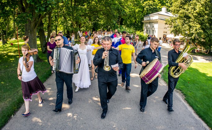 Grupa muzyków gra na instrumentach, idąc parkową aleją.