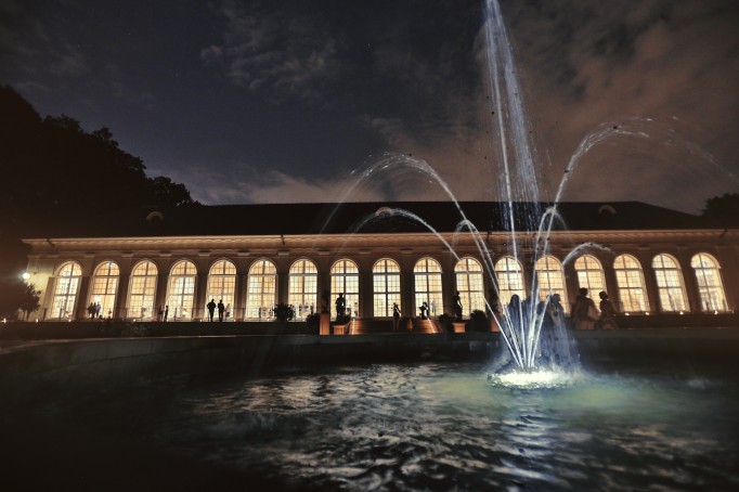 Stara Oranżeria nocą, oświetlona wewnątrz światłem, z przodu budynku widoczna jest tryskająca fontanna.