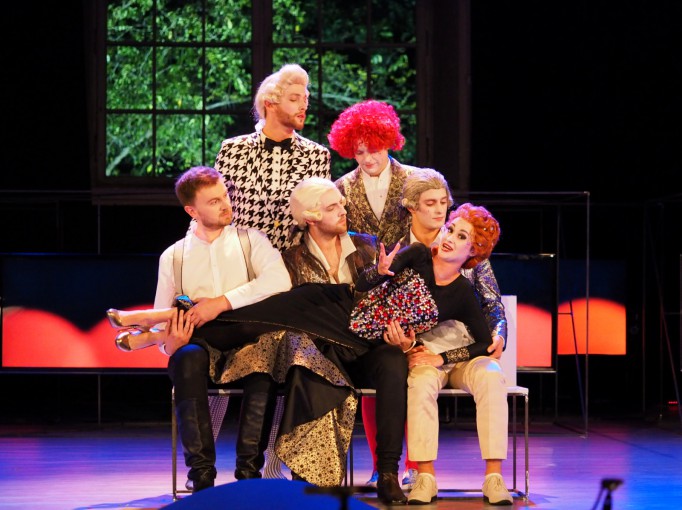 Aktorzy na scenie w kolorowych perukach, trzech z nich siedzi, trzymając leżącą im na kolanach kobietę, dwóch stoi z tyłu.
