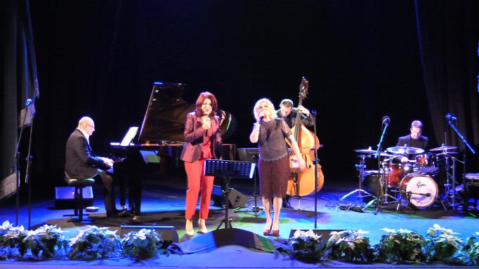 Dwie kobiety stoją na scenie przed mikrofonem i śpiewają, jeden mężczyzna gra na fortepianie, drugi na wiolonczeli.