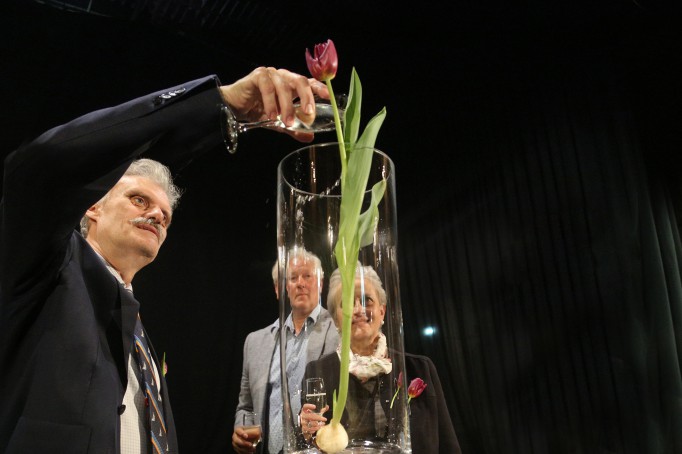 Chrzest tulipana. Do wazonu, w którym stoi kwiat mężczyzna wlewa szampana z kieliszka.