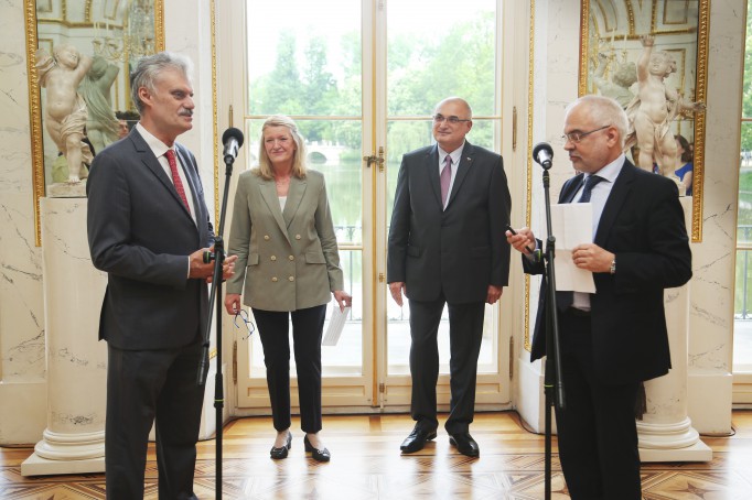 Mężczyzna stojący w sali przed mikrofonem, z tyłu stoi kobieta i mężczyzna.