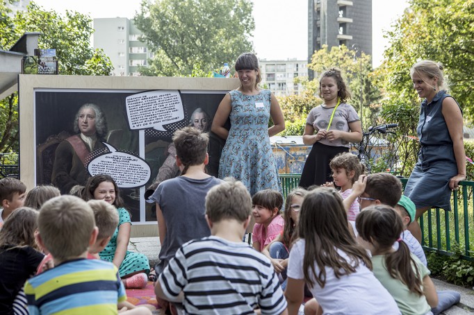 Grupa dzieci siedzi i słucha dwóch kobiet, które stoją na tle obrazu.