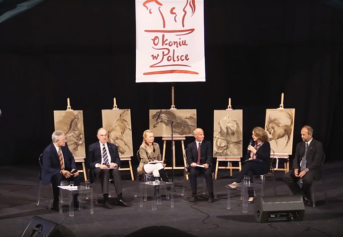 Grupa pięciu osób siedzi na krzesłach ustawionych na scenie i rozmawia, w tle za nimi ustawione są na sztalugach obrazy koni.