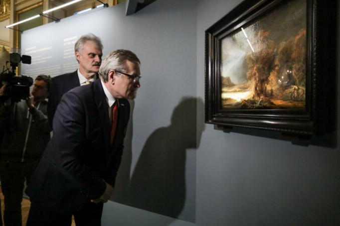 Obraz Rembrandta "Krajobraz z miłosiernym Samarytaninem" wisi na ścianie, przed nim stoi dwóch mężczyzn, którzy go oglądają.