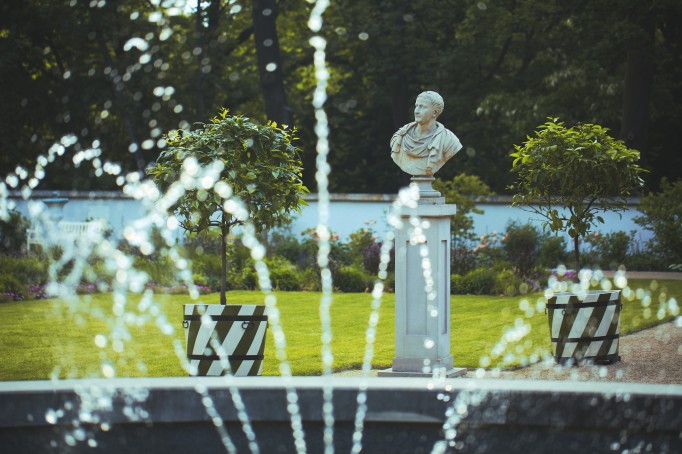 Fragment tryskającej wodą fontanny, w tle egzotyczne drzewka w donicach i marmurowe popiersie, ustawione na cokole.