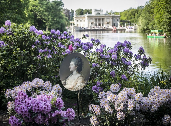Widok na Pałac na Wyspie wśród wody i zieleni. Na pierwszym planie pośród fioletowych kwiatów wetknięta została reprodukacja portretu z kolekcji Stanisława Augusta. 