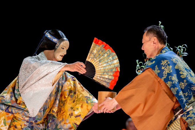 Japoński aktor w masce na twarzy, z wachlarzem w ręku podaje aktorowi w kimonie drewnianą miskę.