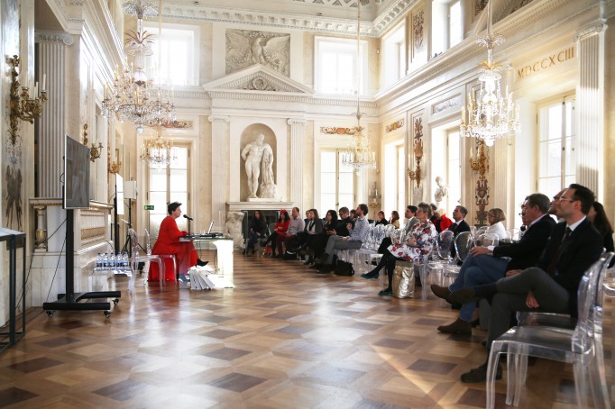 Konferencja w Pałacu na Wyspie. Uczestnicy siedzą na krzesłach, na przeciwko nich siedzi prelegent - kobieta ubrana w długą czerwoną sukienkę.