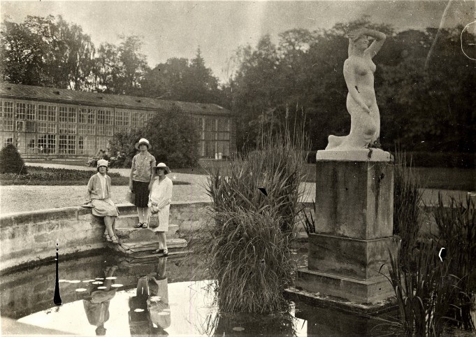 Rok 1927. Trzy kobiety przy sadzawce na tle budynku Starej Oranżerii w Łazienkach Królewskich. Obok przy sadzawce znajduje się rzeźba na postumencie przedstawiająca klęczącą, nagą kobietę.