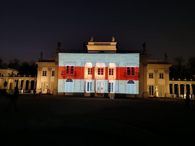 Pałac na Wyspie nocą, oświetlony iluminacją z flagą w barwach historycznej, biało-czerwono-białej flagi Białorusi.