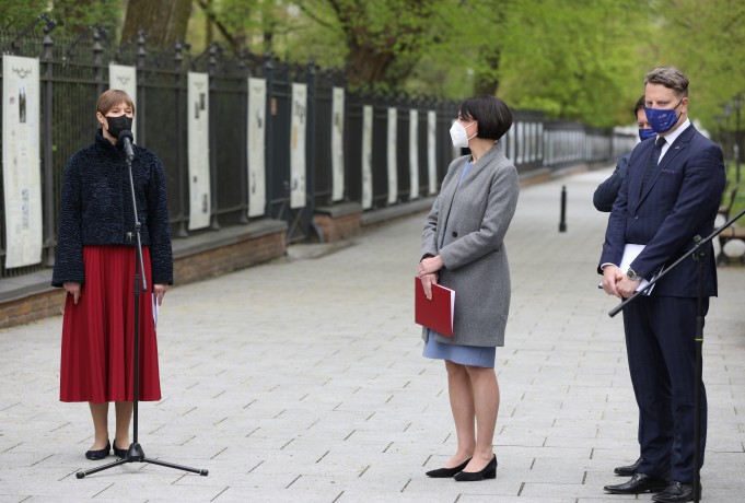 Kobieta w czarnej kurtce, czerwonej spódnicy i maseczce na twarzy stoi przed mikrofonem, na tle plansz w Galerii Plenerowej, z boku obok stoi kobieta i dwóch mężczyzn.