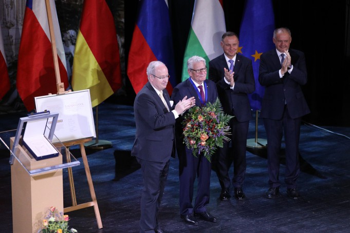 Czterech mężczyzn w ciemnych garniturach stoi na teatralnej scenie. Trzech z nich klaszcze, drugi z lewej trzyma duży bukiet kwiatów.