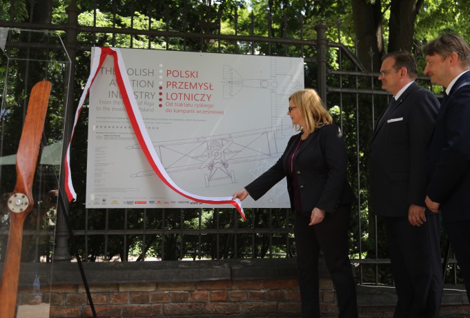 Kobieta zdejmuje biało-czerwoną wstążkę z tablicy, obok niej stoi dwóch mężczyzn w garniturach. 