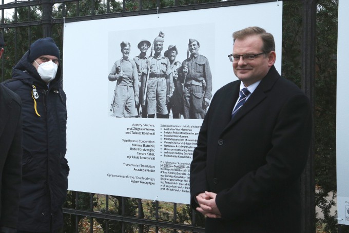 Dwóch mężczyzn stoi przy planszy wystawowej wiszącej na płocie. Plansza przedstawia grupę żołnierzy, trzymających karabiny wiszące na ich ramionach.