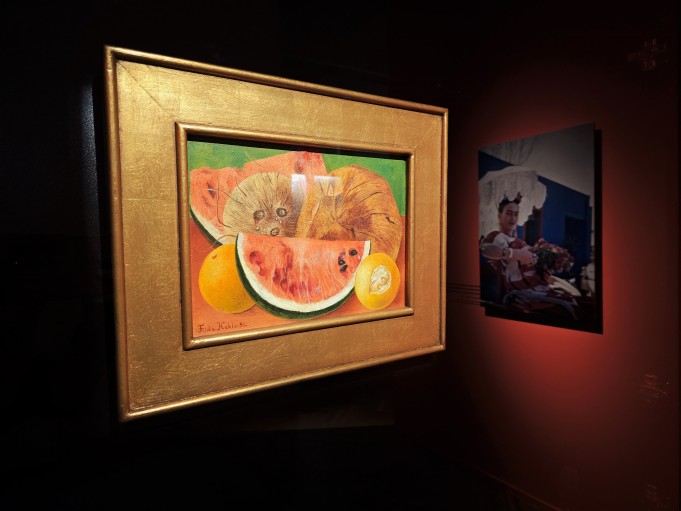 Obraz wiszący na ścianie, który przedstawia martwą naturę. Na obrazie widoczne są dwie pomarańcze, między nimi kawałek arbuza, powyżej kokosy. 