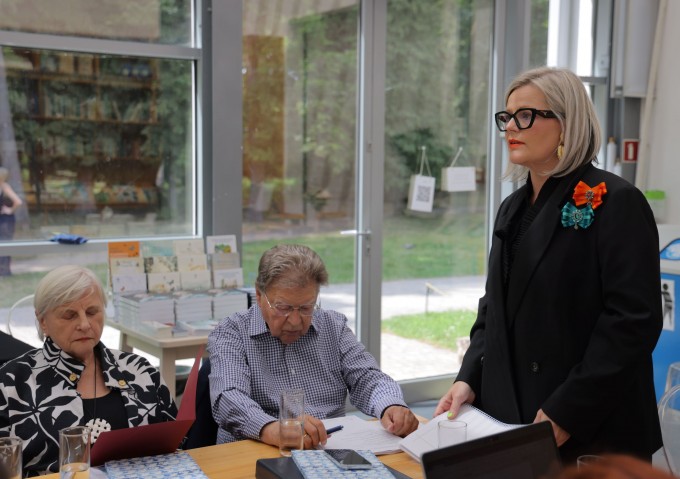 Kobieta w czarnym stroju stoi przy stole, obok, po jej prawej stronie, siedzą przy stole starsza kobieta i mężczyzna.