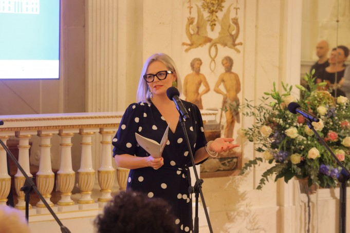 Kobieta w sukience stoi przed mikrofonem i przemawia. 