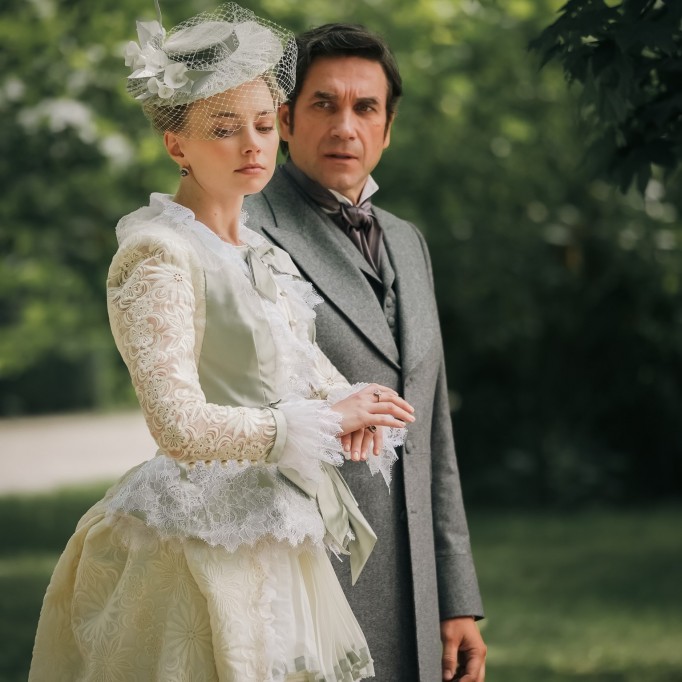 Kobieta w długiej, białej, koronkowej sukni i kapeluszu spaceruje obok mężczyzny w garniturze na tle parku.