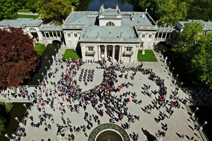 Widok z lotu ptaka na Pałac na Wyspie w otoczeniu wody i zieleni. Na tarasie przed Pałacem widać tłum ludzi.