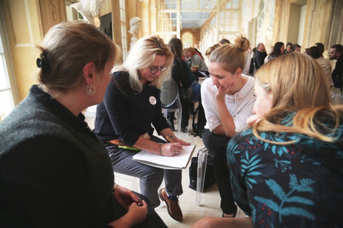 Grupa ludzi siedząca w pomieszczeniu. Kobieta widoczna na pierwszym planie na kolanach trzyma kartkę papieru, na której pisze. 