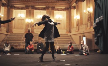 Scena w teatrze, na której siedzą ludzie, jedna z kobiet tańczy, unoszcząc ręce.
