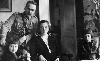Zdjęcie marszałka Piłsudskiego z rodziną. Marszałek, ubrany w mundur, stoi pośrodku, opierając się lewą ręką o fotel, na którym siedzi jego żona. Po lewej i po prawej stronie siedzą jego dwie córki. 