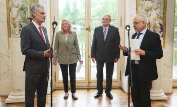 Mężczyzna stojący w sali przed mikrofonem, z tyłu stoi kobieta i mężczyzna.