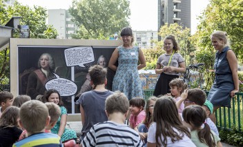 Grupa dzieci siedzi i słucha dwóch kobiet, które stoją na tle obrazu.
