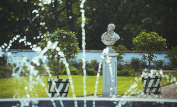 Fragment tryskającej wodą fontanny, w tle egzotyczne drzewka w donicach i marmurowe popiersie, ustawione na cokole.
