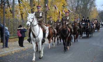 Kawalerzyści w mundurach jadą konno, z tyłu z nimi jadą dorożki. Ludzie, którzy stoją wzdłuż ulicy, robią im zdjęcia.