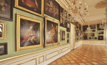 Galeria Obrazów w Pałacu na Wyspie. Obrazy z kolekcji króla Stanisława Augusta w złotych ramach wiszą na jasnozielonej ścianie. Z sufitu zwisają trzy kryształowe żyrandole.