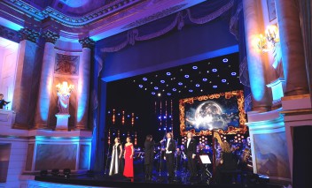 Koncert w Teatrze Królewskim. Na scenie znajduje się orkiestra i soliści, widać fragment widowni. 