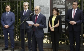 Trzech mężczyzn z lewej, kobieta i mężczyzna z prawej strony stoją pod ogrodzeniem z planszami wystawowymi, mężczyzna trzeci od lewej przemawia do mikrofonu, trzymając kartę w ręku. 