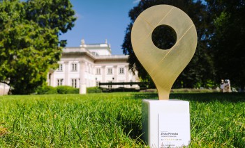 Statuetka Złota Pinezka stojąca na trawie, na tle Pałacu na Wyspie.