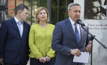 Mężczyzna w garniturze stoi przed mikrofonem, z tyłu za nim stoi kobieta z mężczyzną, widoczny jest też fragment planszy wystawowej wiszącej na płocie.