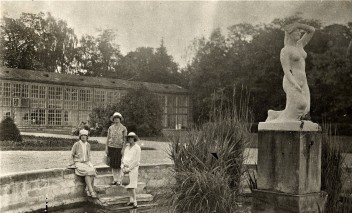 Rok 1927. Trzy kobiety przy sadzawce na tle budynku Starej Oranżerii w Łazienkach Królewskich. Obok przy sadzawce znajduje się rzeźba na postumencie przedstawiająca klęczącą, nagą kobietę.
