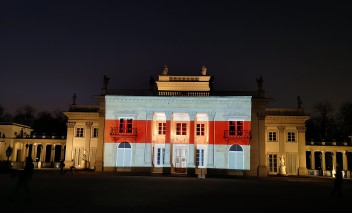 Pałac na Wyspie nocą, oświetlony iluminacją z flagą w barwach historycznej, biało-czerwono-białej flagi Białorusi.