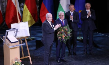Czterech mężczyzn w ciemnych garniturach stoi na teatralnej scenie. Trzech z nich klaszcze, drugi z lewej trzyma duży bukiet kwiatów.