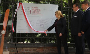 Kobieta zdejmuje biało-czerwoną wstążkę z tablicy, obok niej stoi dwóch mężczyzn w garniturach. 