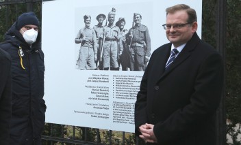 Dwóch mężczyzn stoi przy planszy wystawowej wiszącej na płocie. Plansza przedstawia grupę żołnierzy, trzymających karabiny wiszące na ich ramionach.