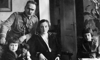 Zdjęcie marszałka Piłsudskiego z rodziną. Marszałek, ubrany w mundur, stoi pośrodku, opierając się lewą ręką o fotel, na którym siedzi jego żona. Po lewej i po prawej stronie siedzą jego dwie córki. 