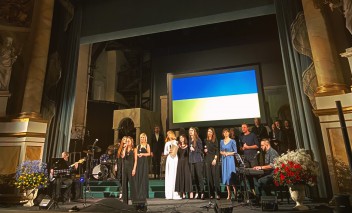 Grupa artystów stoi na scenie Teatru Królewskiego i śpiewa. Nad artystami wisi ukraińska flaga w niebiesko-żółtych barwach.
