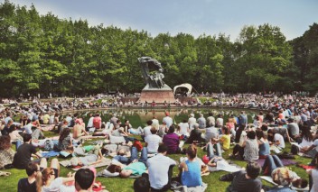 Ludzie siedzą na trawie wokół Pomnika Chopina w Łazienkach Królewskich. 