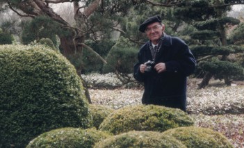 Mężczyzna w czarnej czapce i płaszczu stoi w ogrodzie wśród kilku krzewów mających kształt kuli. 