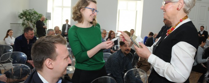 Grupa osób siedzi na sali, wśród zebranych jedna z kobiet wręcza drugiej swoją wizytówkę.