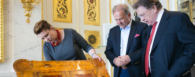 Stolik do gry w karty króla Stanisława Augusta. Przy meblu stoi dwóch mężczyzn i kobieta, która go ogląda.