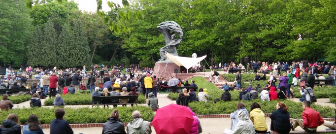 Ludzie siedzą na trawie wokół Pomnika Chopina i słuchają koncertu.