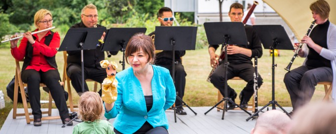 Muzycy podczas koncertu grają na intrumentach, z przodu kuca kobieta, która pokazuje dzieciom maskotkę.