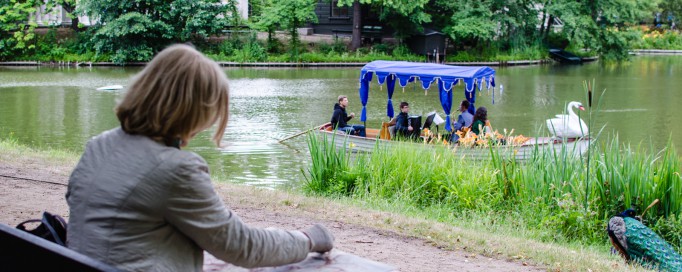 Kobieta siedzi w parku i maluje, przed nią znajduje się rzeka, po której płynie gondola.